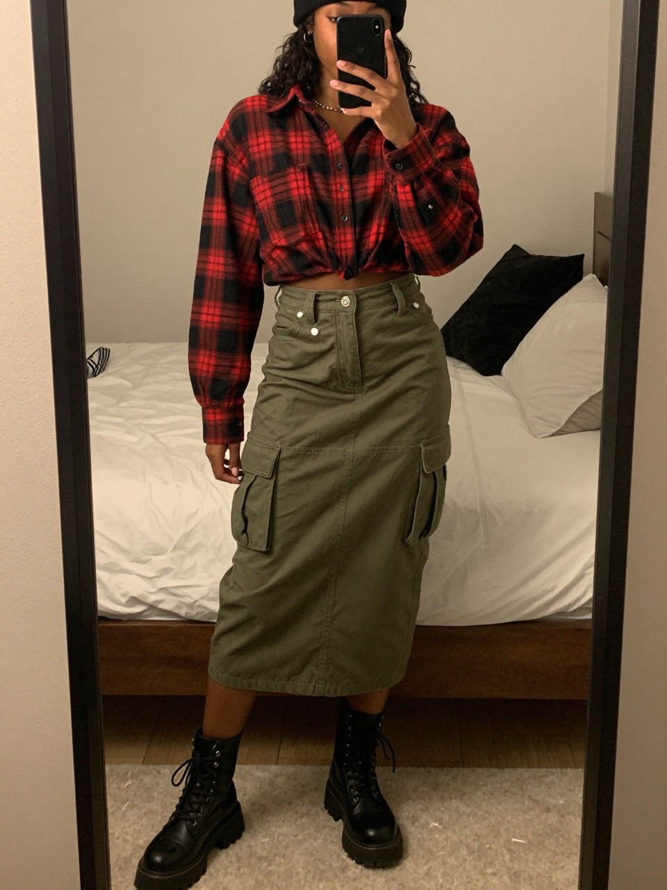 iPhone mirror selfie of a black woman wearing a red and black plaid flannel shirt tucked into an olive green cargo midi skirt with black chunky combat boots and a black beanie, standing in a relaxed grunge-inspired pose, cropped at NECK so face doesn't show, in a feminine-themed bedroom, warm moody lighting, iPhone photo quality.