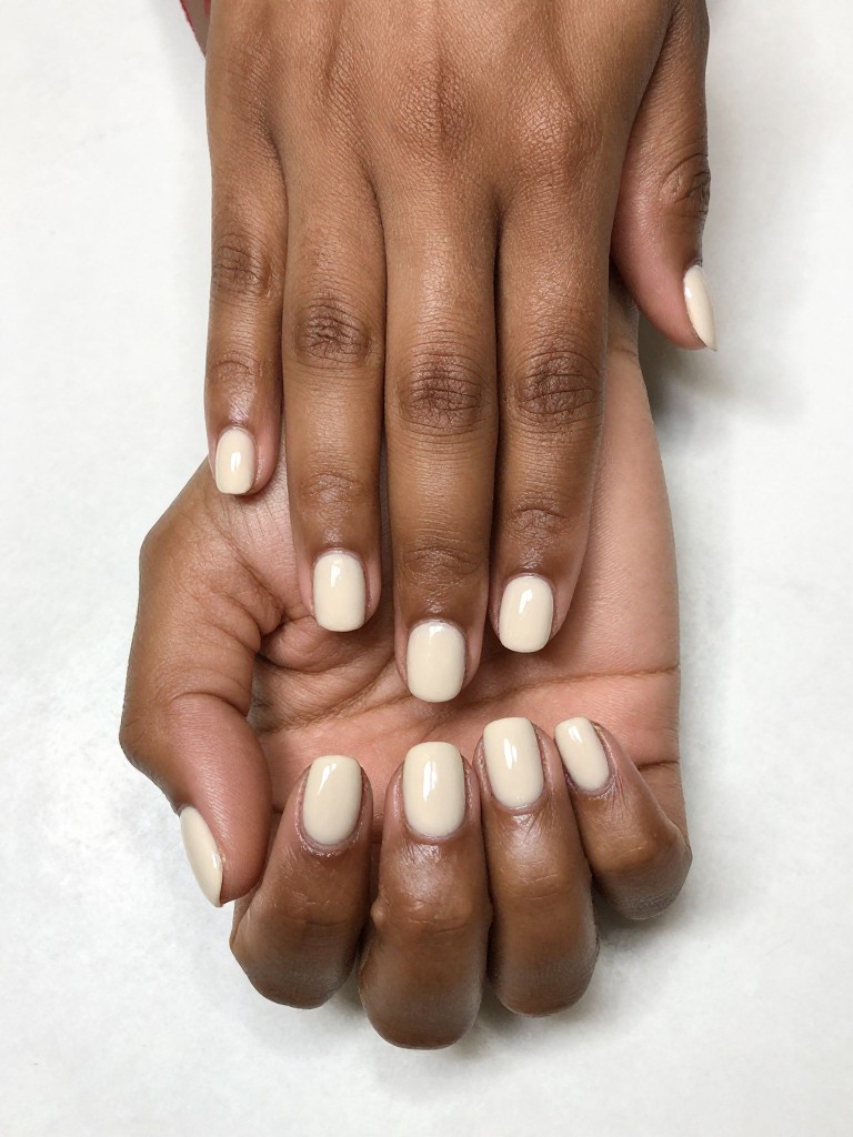 Overhead, top-down view of black feminine hands with very short, rounded nails resting palms down on a smooth, white stone surface. The nails are coated in a creamy, neutral nude polish with a flawless, mirror-like glossy finish. The lighting is bright and even, emphasizing the healthy, clean look.