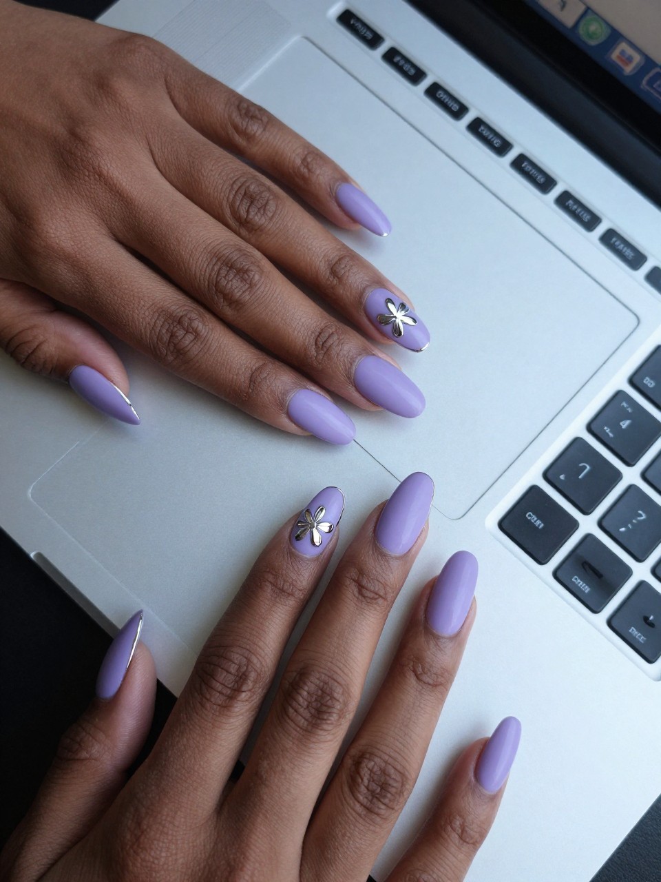 Top-down view of black feminine hand with coffin nails on a sleek silver laptop. The nails are a frosty matte lilac. A crisp, mirror-like chrome outline of a simple flower is painted on each nail. The lighting is cool and tech-inspired.
