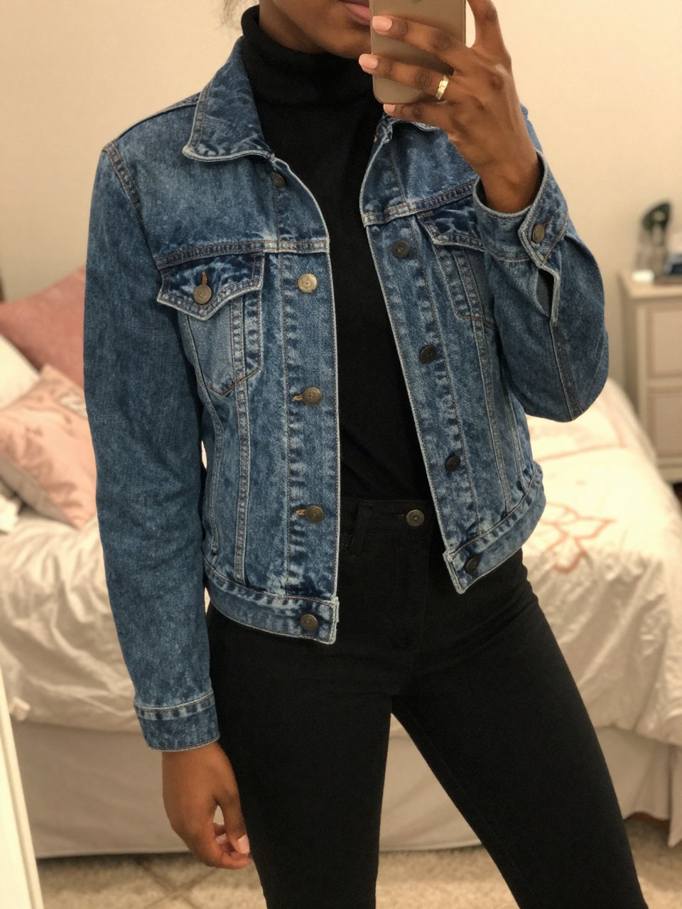 iPhone mirror selfie of a black woman in a jean jacket over a black turtleneck with black pants, angled slightly, cropped at NECK so face doesn't show, in a feminine-themed bedroom with warm moody lighting, iPhone photo quality.