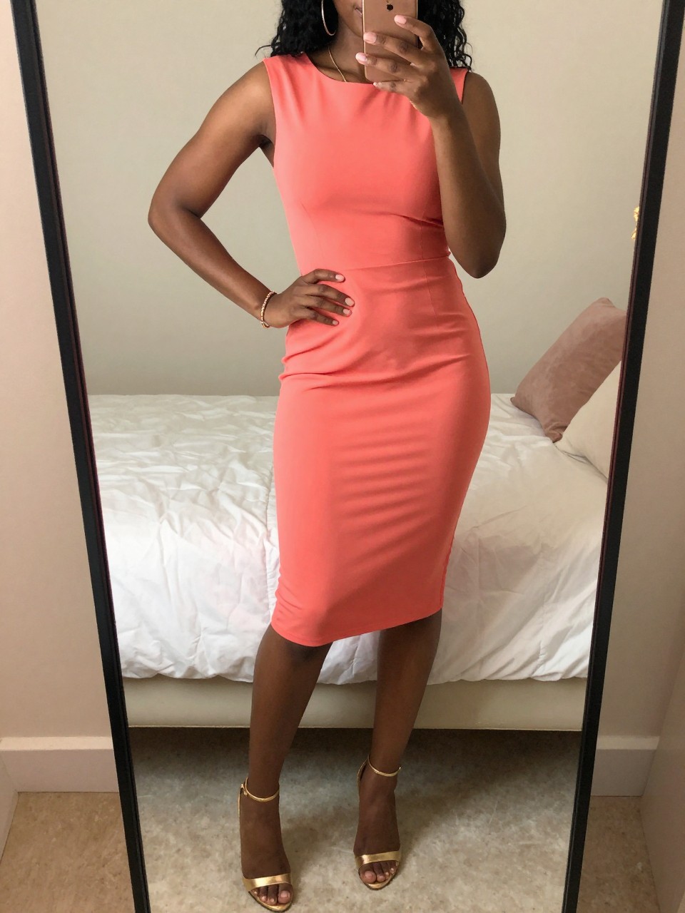 iPhone mirror selfie of a Black woman wearing a coral dress with gold heels and accessories, standing with one hand on her hip and phone in the other, cropped at NECK so face doesn't show, in a bright feminine-themed bedroom, warm moody lighting, iPhone photo quality.