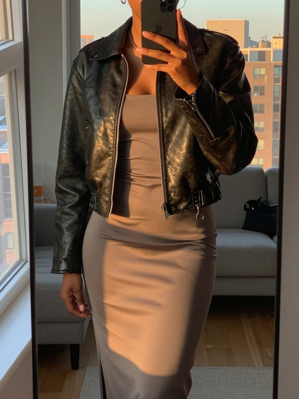 iPhone mirror selfie of a black woman in a leather jacket and slip skirt with Timbs, one hand slightly raised, cropped at NECK so face doesn't show, in a city apartment during evening golden hour, iPhone photo quality.