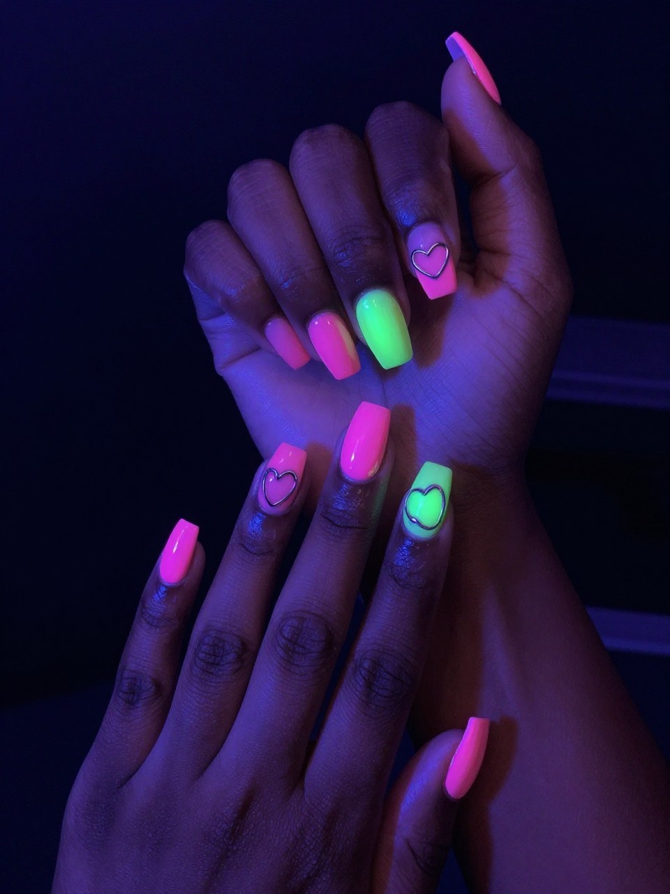 Black feminine hands in a darkened room with UV light. The nails have a bright neon pink or green glow. The outlines of hearts in chrome reflect the ambient light, creating a double-layered, electrifying effect. Perfect for a night out.