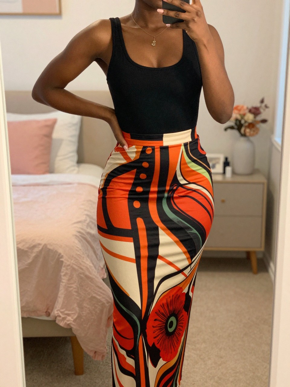 iPhone mirror selfie of a black woman wearing a black tank top and a bold printed maxi skirt, posed with one hand on her hip, cropped at NECK so face doesn't show, in a bright feminine-themed bedroom, warm moody lighting, iPhone photo quality.
