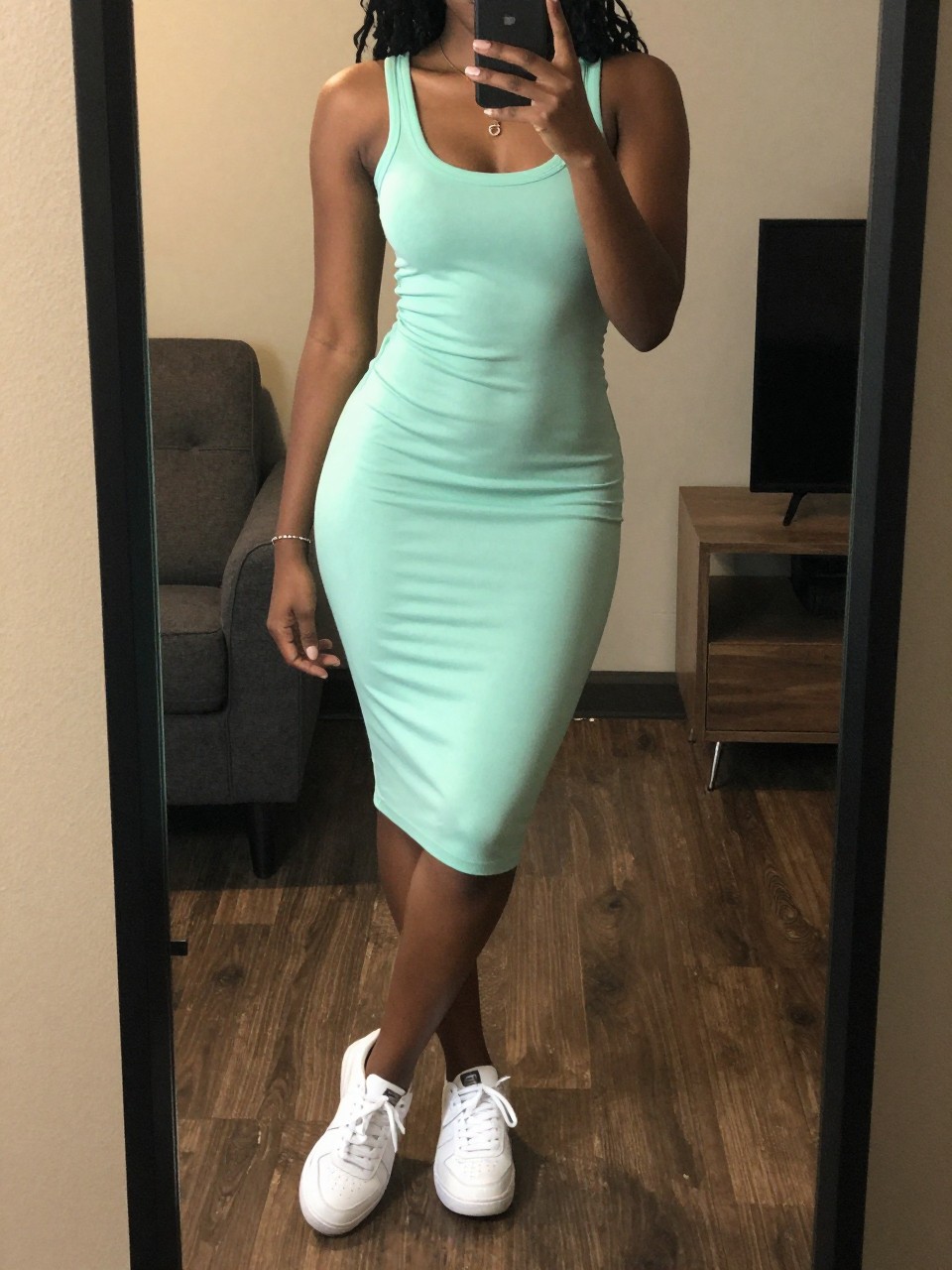 iPhone mirror selfie of a Black woman wearing a mint green dress with white sneakers, standing casually with one foot forward and phone in hand, cropped at NECK so face doesn't show, in a city apartment, warm moody lighting, iPhone photo quality.