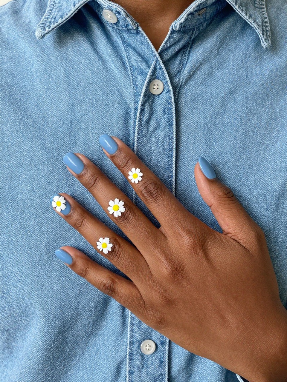 Top-down view of black feminine hands with short nails resting on a light blue denim shirt. The nails are a matte powder blue. White daisies with yellow centers are painted with tiny black “stitch” marks around them. The vibe is casual, cute, and handmade.