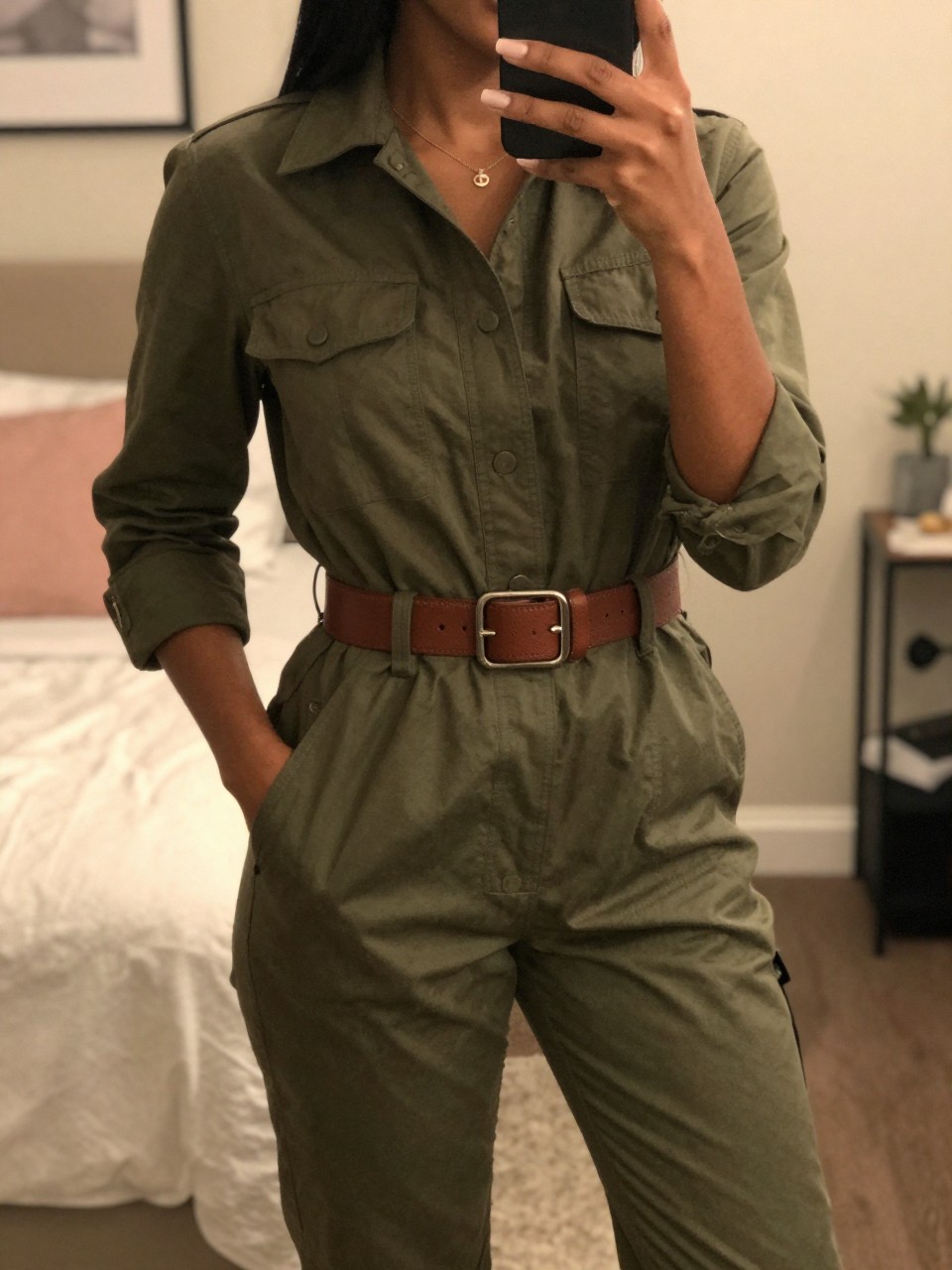 iPhone mirror selfie of a black woman wearing an olive utility jumpsuit with a cognac belt, posed with one hand in her pocket, cropped at NECK so face doesn't show, in a feminine-themed bedroom, warm moody lighting, iPhone photo quality.