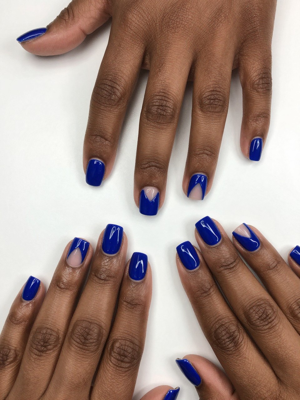 Overhead view of black feminine hand with neat, short nails on a white desk. The nails are glossy cobalt blue. A clean, heart-shaped window of bare nail is exposed in the center of each. The design is striking and architectural.