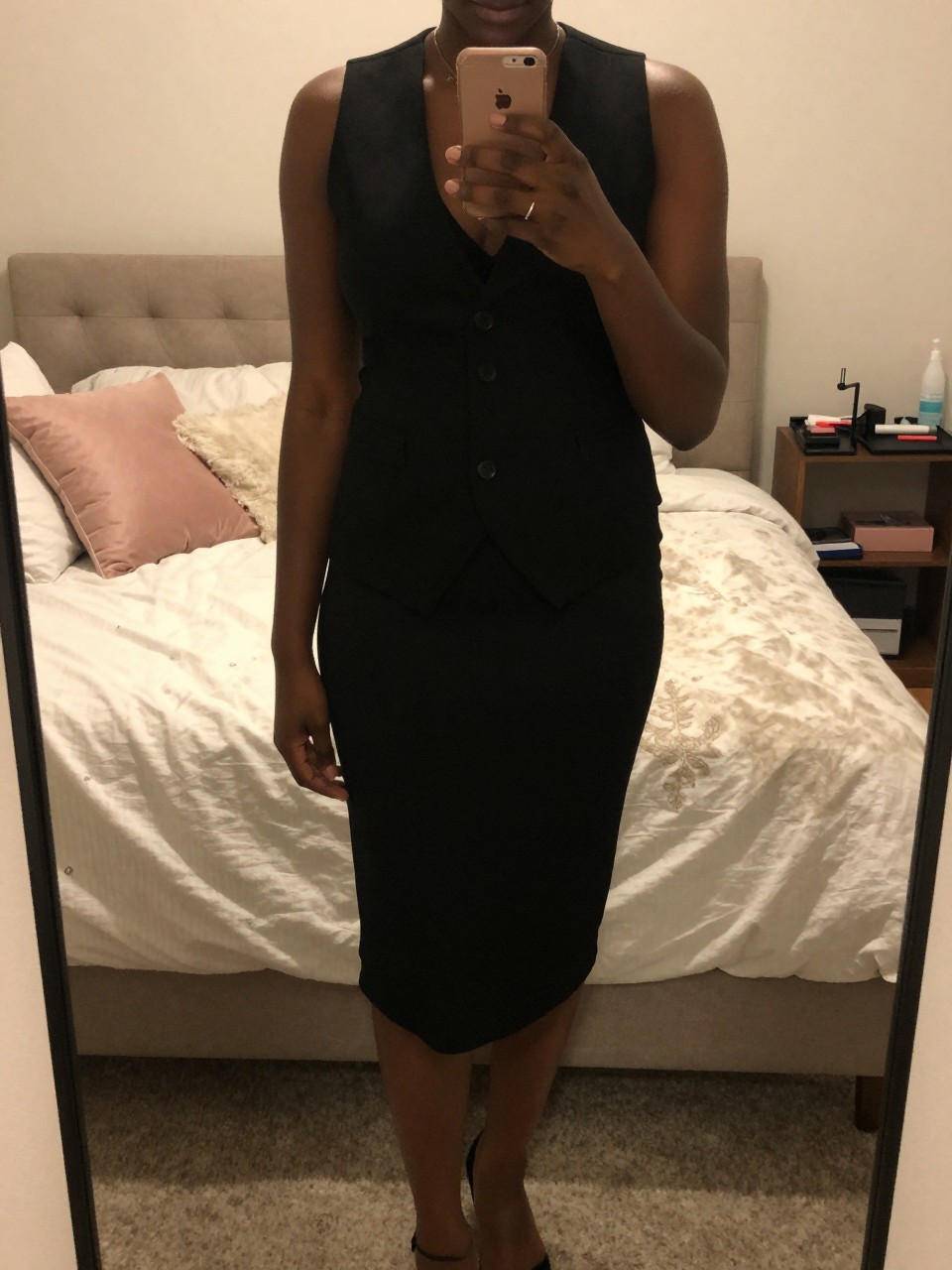 iPhone mirror selfie of a black woman wearing a black vest over a slip dress, standing with her phone low and one foot forward, cropped at NECK so face doesn't show, in a feminine-themed bedroom, warm moody lighting, iPhone photo quality.