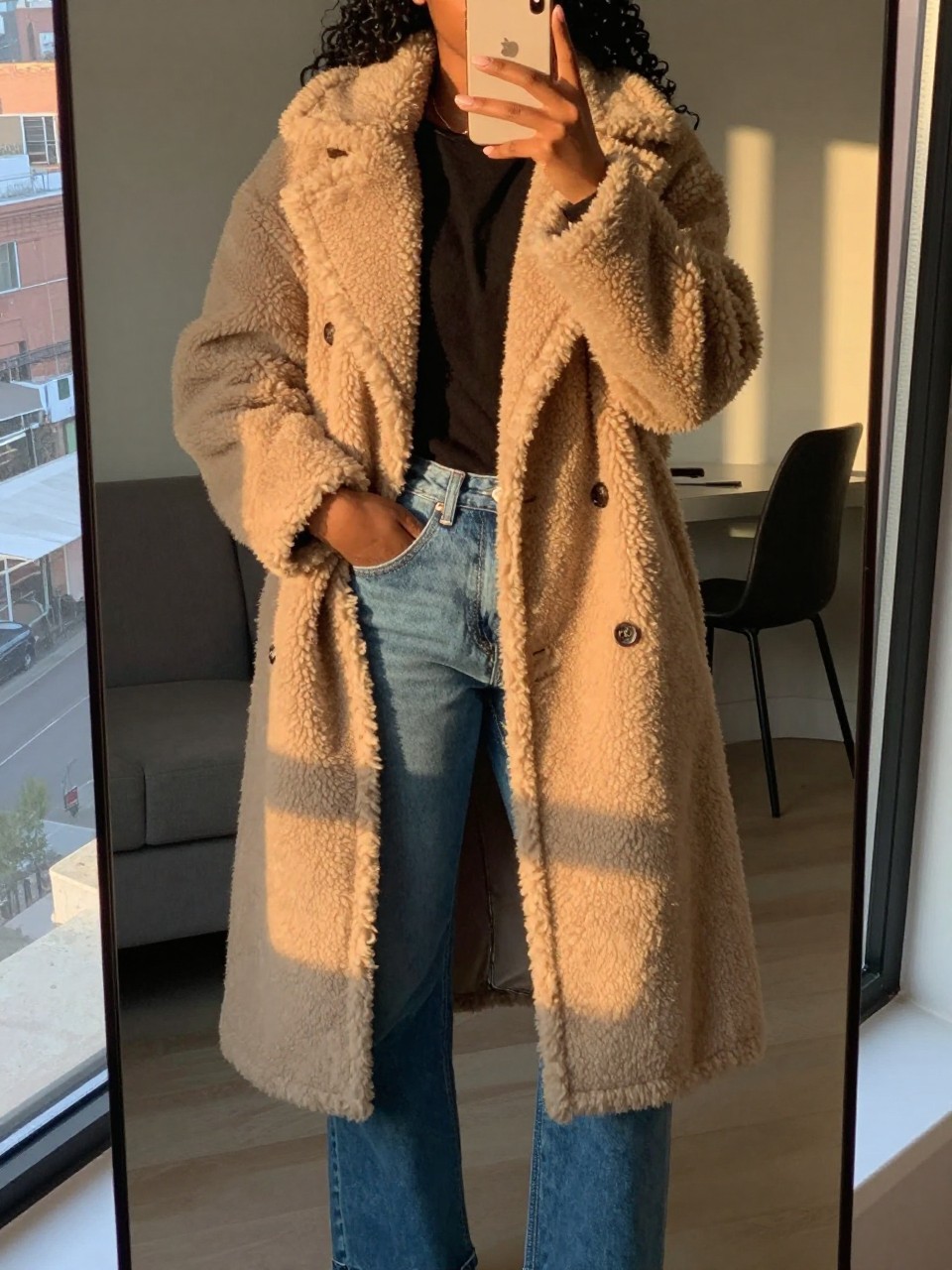 iPhone mirror selfie of a black woman in a teddy coat and straight jeans with Timbs, hand in coat pocket, cropped at NECK so face doesn't show, in a city apartment during evening golden hour, iPhone photo quality.