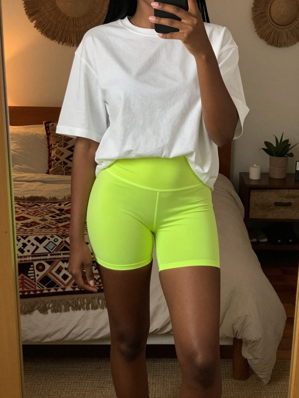 iPhone mirror selfie of a black woman in an oversized tee and neon bike shorts, one foot forward, cropped at NECK so face doesn't show, in a bohemian-themed bedroom with warm moody lighting, iPhone photo quality.