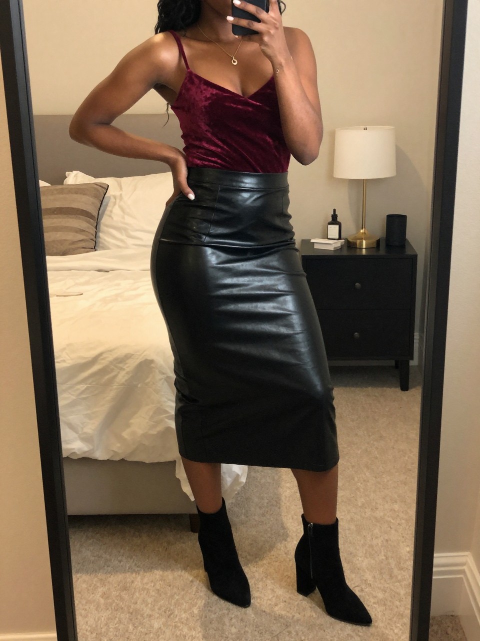 iPhone mirror selfie of a black woman wearing a burgundy velvet camisole tucked into a black faux leather midi skirt with black suede block-heel ankle boots, one hand on her hip, cropped at NECK so face doesn't show, in a feminine-themed bedroom, warm moody lighting, iPhone photo quality.