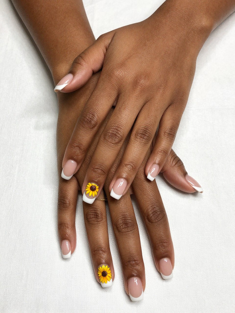 Overhead view of black feminine hands with classic French manicures, resting on a white tablecloth. All nails have a white tip. One ring finger features a single, detailed yellow sunflower with a brown center painted on the nail bed. The contrast is elegant and bold.