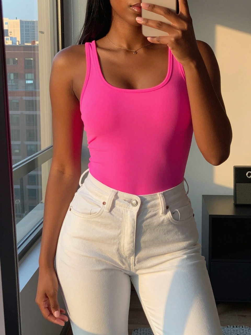mirror selfie of a black woman in a neon pink top and white jeans, hand slightly raised, cropped at NECK so face doesn't show, in a city apartment during evening golden hour, iPhone photo quality.