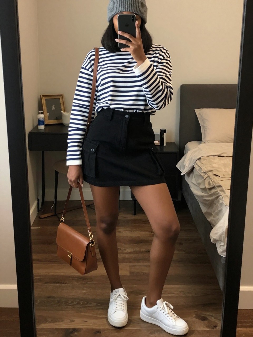 iPhone mirror selfie of a black woman wearing a navy and white striped long-sleeve shirt tucked into a black cargo mini skirt with white sneakers and a grey knit beanie, holding a leather crossbody bag, standing in a relaxed city-ready pose, cropped at NECK so face doesn't show, in a feminine-themed bedroom, warm moody lighting, iPhone photo quality.