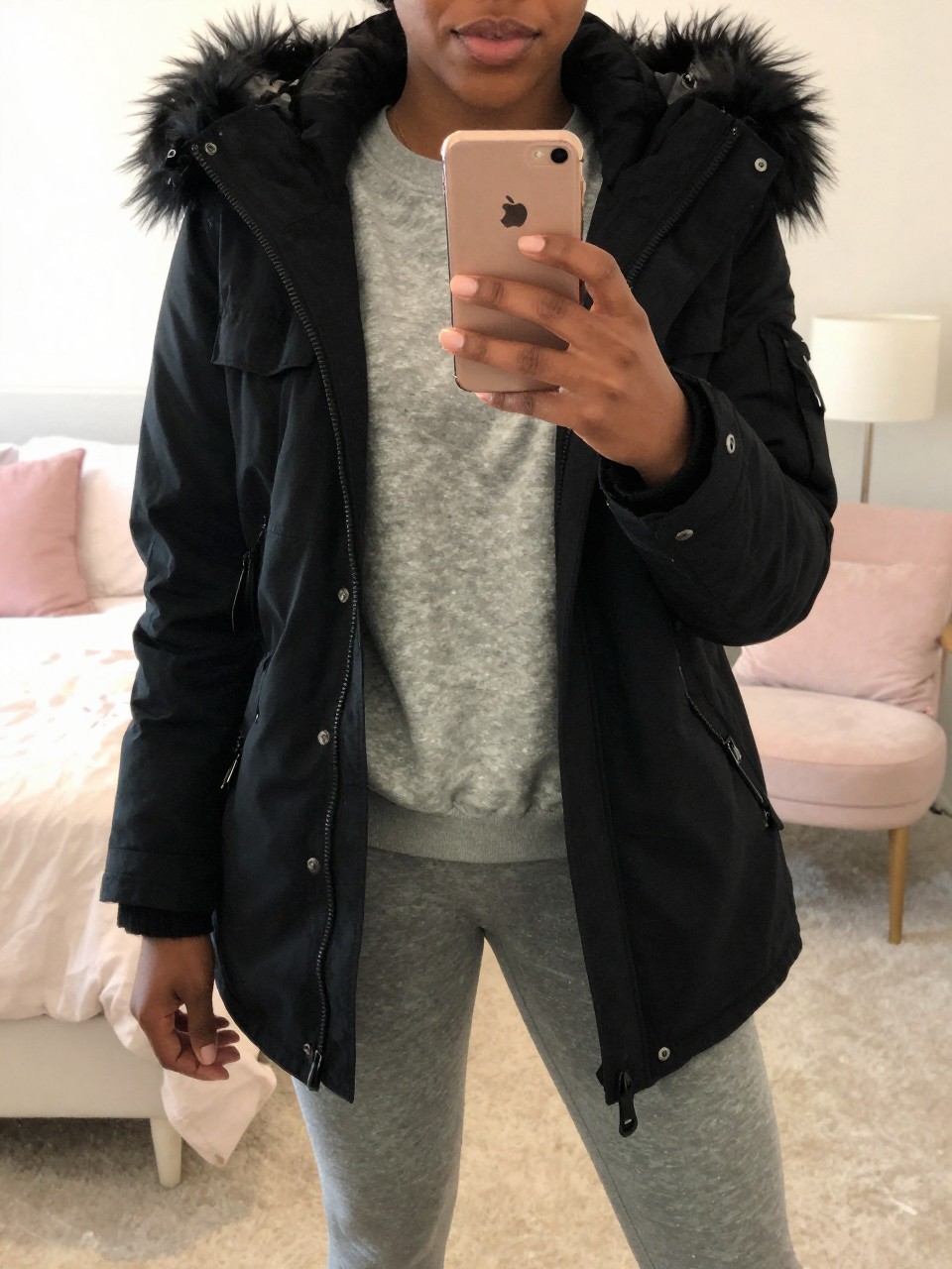 iPhone mirror selfie of a Black woman wearing a black parka with faux fur trim over a gray sweatshirt and leggings, standing with her phone in front, cropped at NECK so face doesn't show, in a bright feminine-themed bedroom, warm moody lighting, iPhone photo quality.