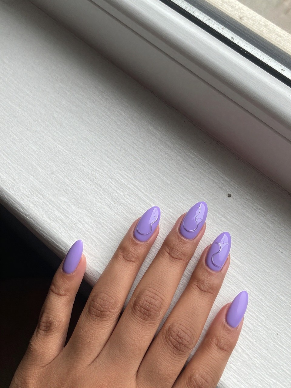 Overhead view of black feminine hands with almond nails on a concrete windowsill. The nails are a matte violet. A single, glossy clear line curves on each nail to form a minimalist flower silhouette. The natural daylight highlights the glossy detail.