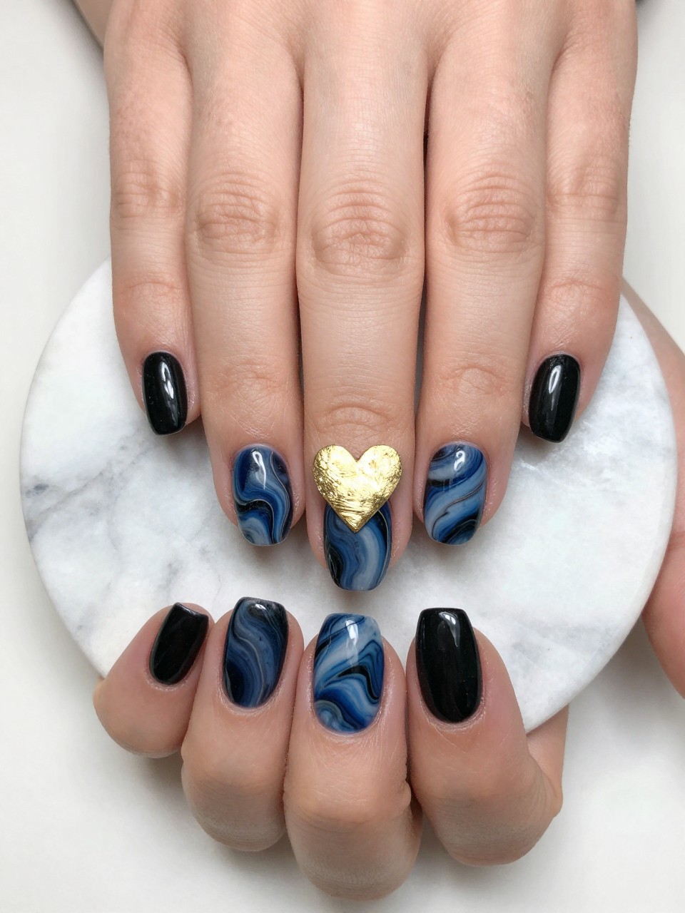 Close-up, top-down view of a black feminine nail with a blue marble swirl. A delicate gold foil heart is placed in the center. The hands rest on a marble coaster, tying the elegant theme together.