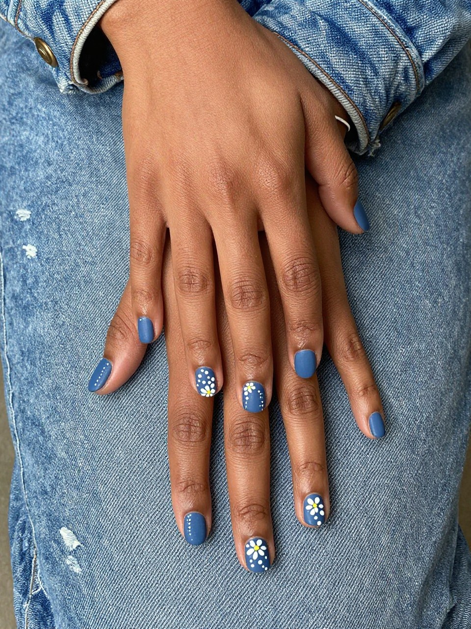 Bird's-eye view of black feminine hands with short, rounded nails resting on a worn denim jacket. The nails are a matte denim blue. Simple white dot flowers with tiny yellow centers are scattered randomly on each nail. The setting is relaxed and everyday.