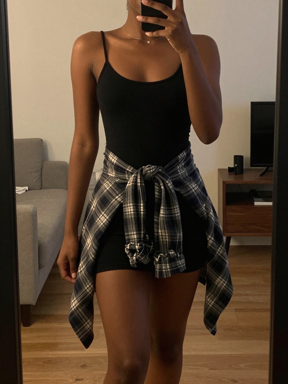 iPhone mirror selfie of a black woman wearing a black slip dress with a flannel tied at her waist and wheat Timbs, posed with one knee slightly bent, cropped at NECK so face doesn't show, in a city apartment with warm moody lighting, iPhone photo quality.