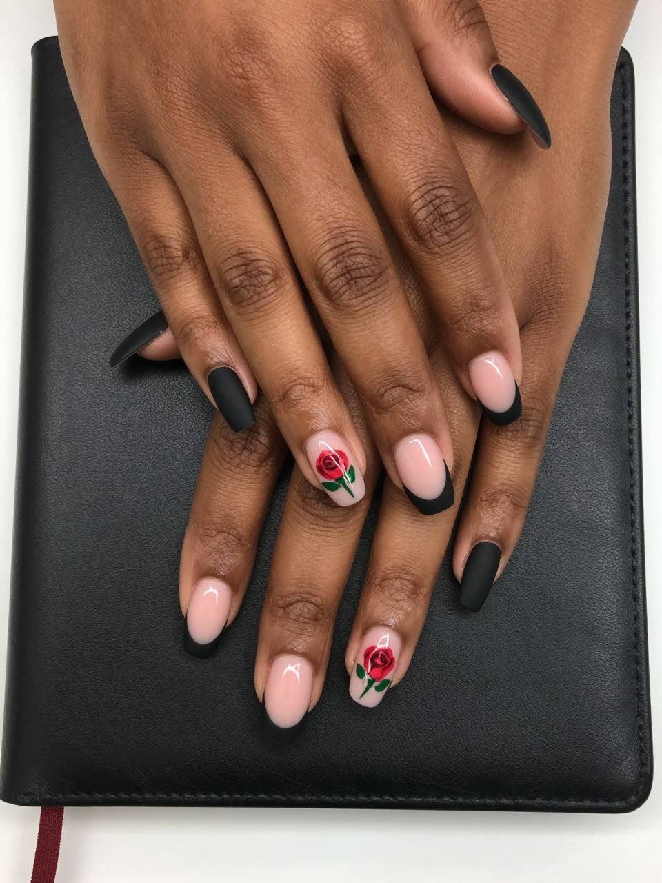 Overhead view of black feminine hands with nails resting on a black leather journal. The French tips are matte black. On the sheer pink base of each nail, a single red rosebud with a green leaf is painted. The mood is bold and romantic.
