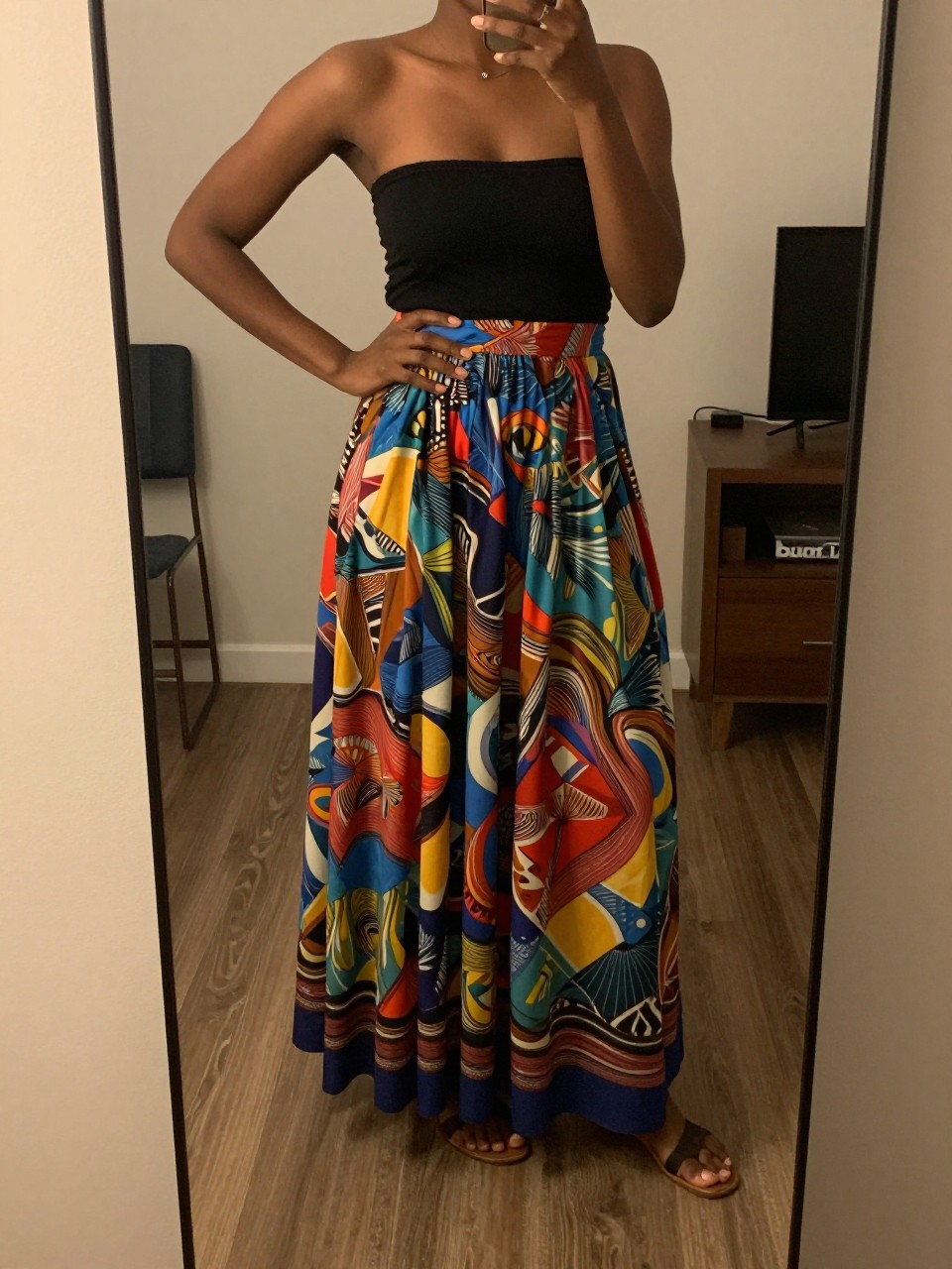 iPhone mirror selfie of a black woman wearing a black bandeau top with a colorful printed flowy maxi skirt and flat leather sandals, one hand on her hip, cropped at NECK so face doesn't show, in a city apartment, warm moody lighting, iPhone photo quality.