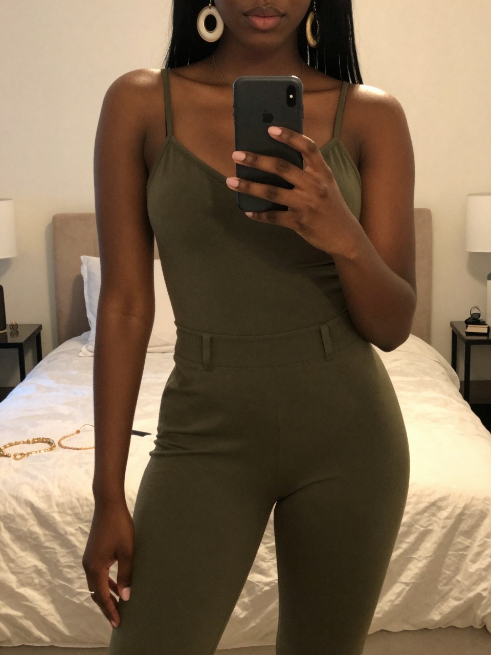 mirror selfie of a black woman in an olive green jumpsuit and statement earrings, holding phone at chest level, cropped at NECK so face doesn't show, in a feminine-themed bedroom with warm moody lighting, iPhone photo quality.