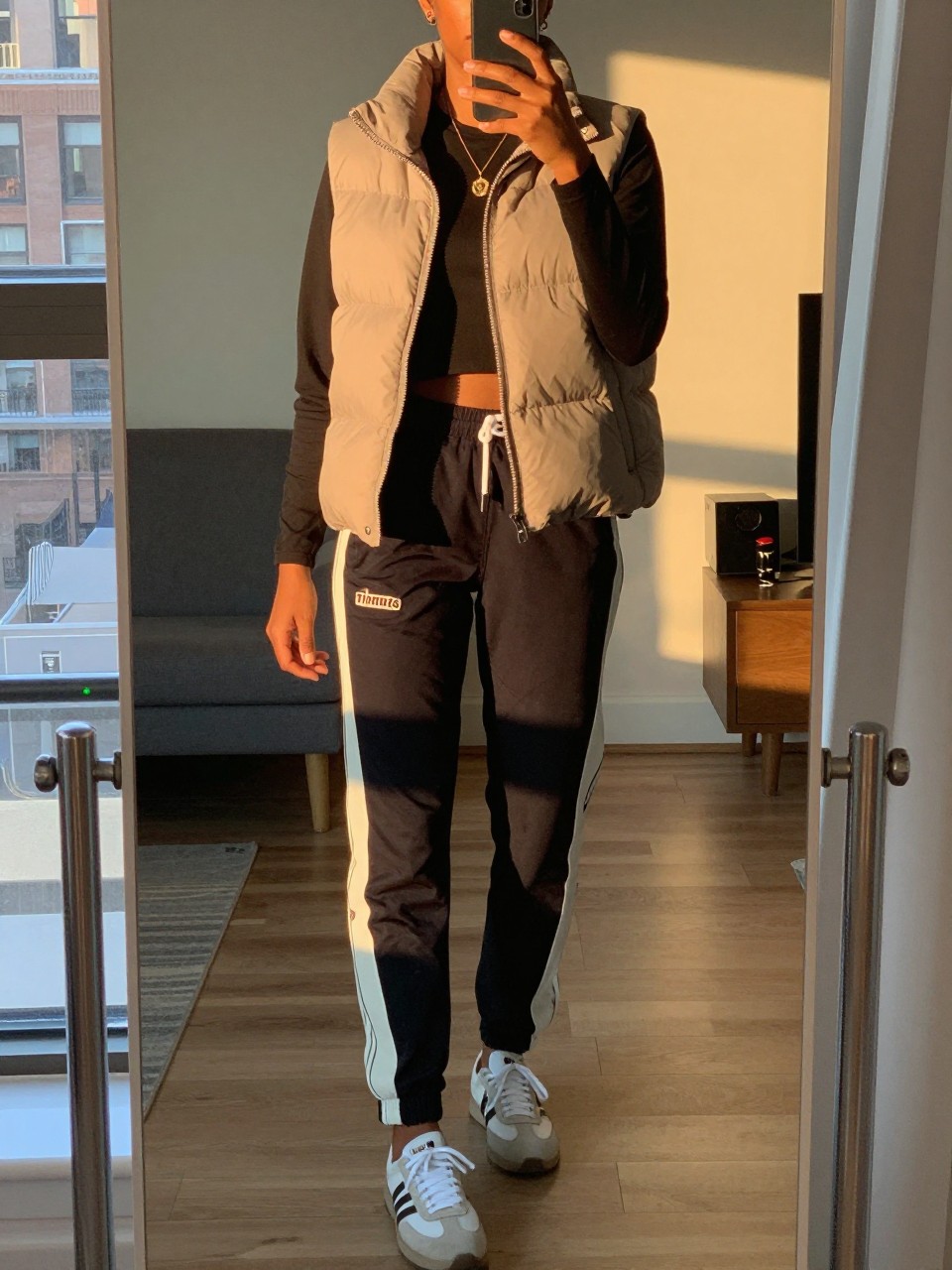 iPhone mirror selfie of a black woman in a puffer vest and track pants with Timbs, one foot forward, cropped at NECK so face doesn't show, in a city apartment during evening golden hour, iPhone photo quality.