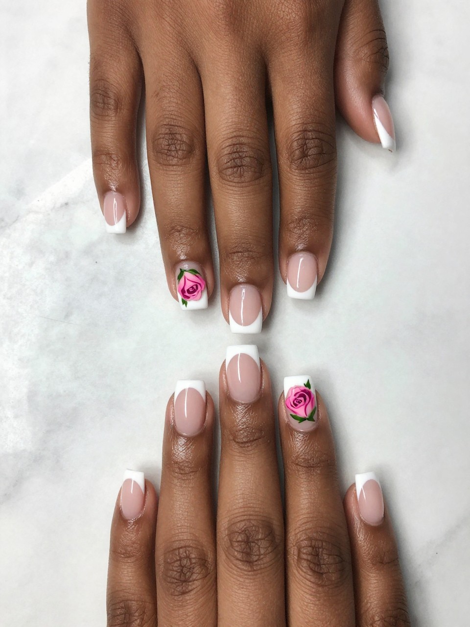 Overhead shot of black feminine hands with medium square nails on a marble surface. The nails feature a crisp white French tip. On the side of each nail, near the cuticle, a single pink rosebud with a green leaf is painted. The composition is asymmetrical and chic.