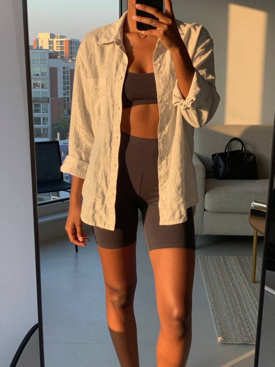 iPhone mirror selfie of a black woman in an open linen shirt over bike shorts, one foot slightly forward, cropped at NECK so face doesn't show, in a city apartment during evening golden hour, iPhone photo quality.