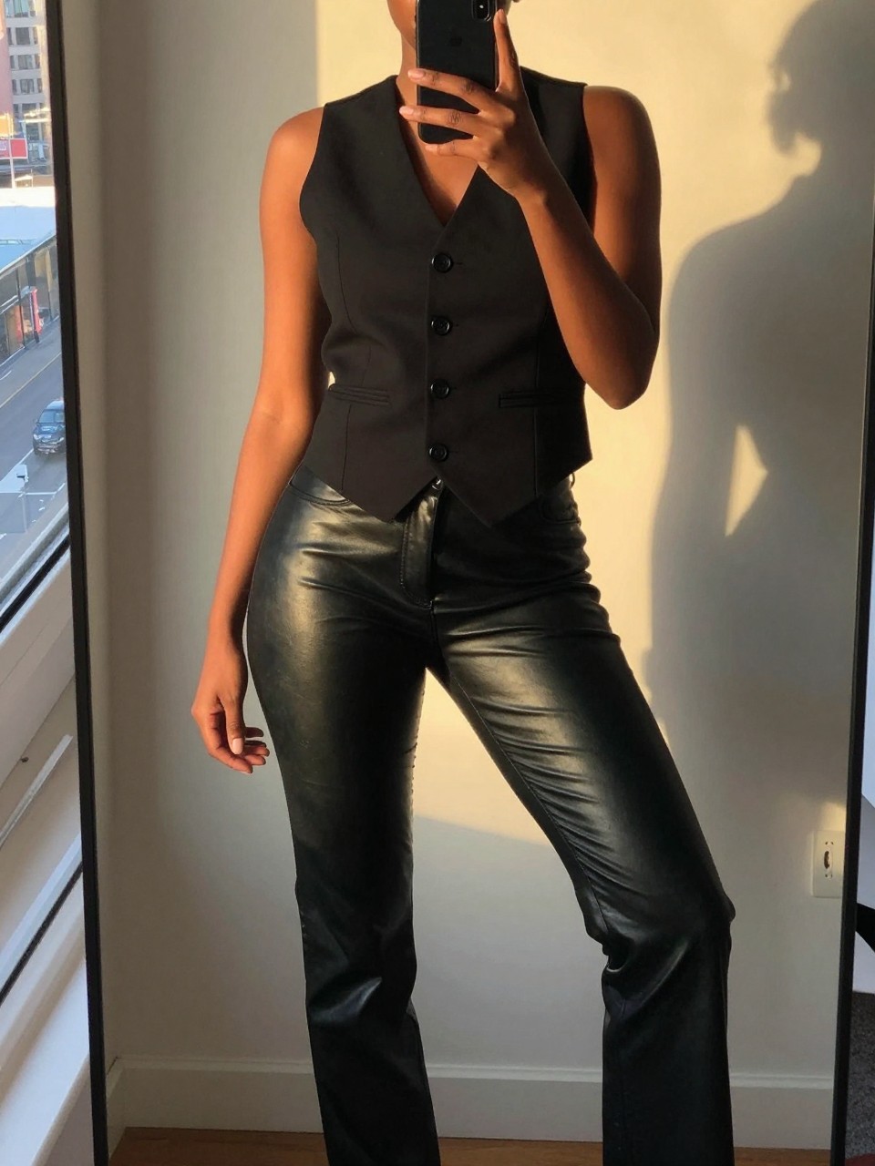 iPhone mirror selfie of a black woman wearing a black vest with leather pants, standing with her phone arm relaxed and one leg slightly bent, cropped at NECK so face doesn't show, in a city apartment, evening golden hour light, iPhone photo quality.