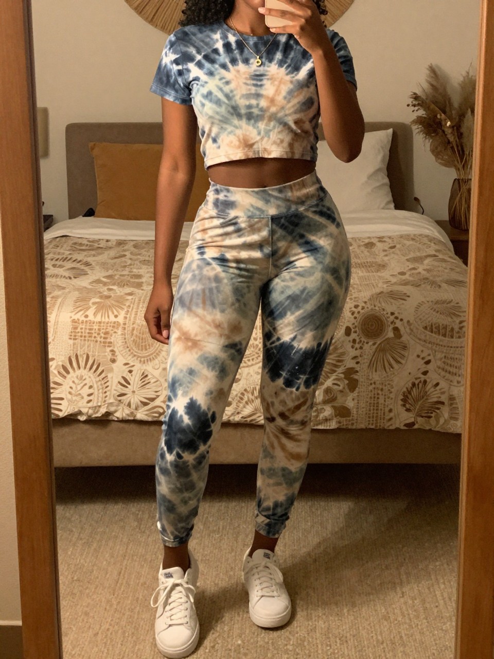 mirror selfie of a black woman in a tie-dye matching set and white sneakers, posed with one foot forward, cropped at NECK so face doesn't show, in a bohemian-themed bedroom with warm moody lighting, iPhone photo quality.
