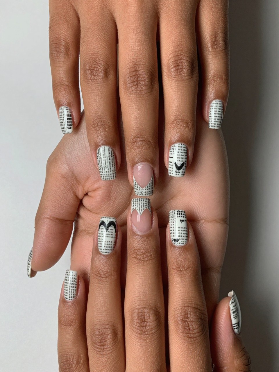 Top-down view of black feminine hands with squared nails. The nails are covered in a fine newspaper print pattern. A perfect heart shape is missing from the print on each nail, showing bare nail underneath. The look is literary and rebellious.