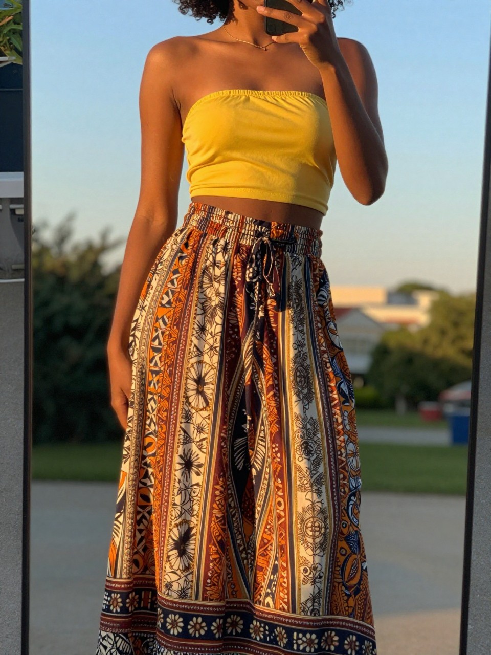 mirror selfie of a black woman in a yellow bandeau top and printed maxi skirt, phone held low to show full skirt, cropped at NECK so face doesn't show, in a sunny outdoor setting during evening golden hour, iPhone photo quality.