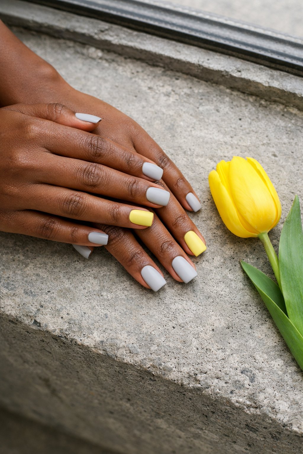 Top-down view of black hands with square nails resting on a concrete windowsill next to a yellow tulip. Most nails are a light matte grey. The ring fingers are painted a soft matte pastel yellow. The composition is minimalist and artsy.
