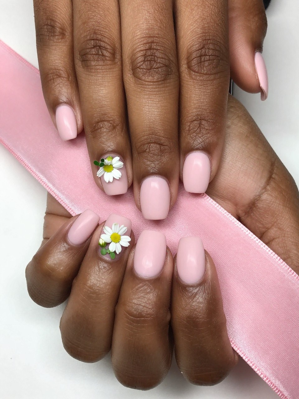 Close-up, top-down view of black feminine hands with short, rounded nails on a pink velvet ribbon. The nails are a matte ballet slipper pink. on the nails is a tiny cluster of closed white daisy buds with micro green leaves sits at the cuticle of each nail. The look is precious and soft. Make it a realistic nail design