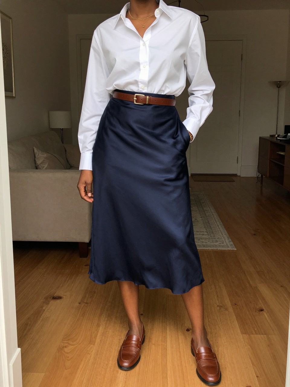 photo of a black woman wearing a white button-down shirt tucked into a navy satin midi skirt with brown leather loafers and a matching belt, one hand in her pocket, cropped at NECK so face doesn't show, in a city apartment, warm moody lighting, iPhone photo quality.