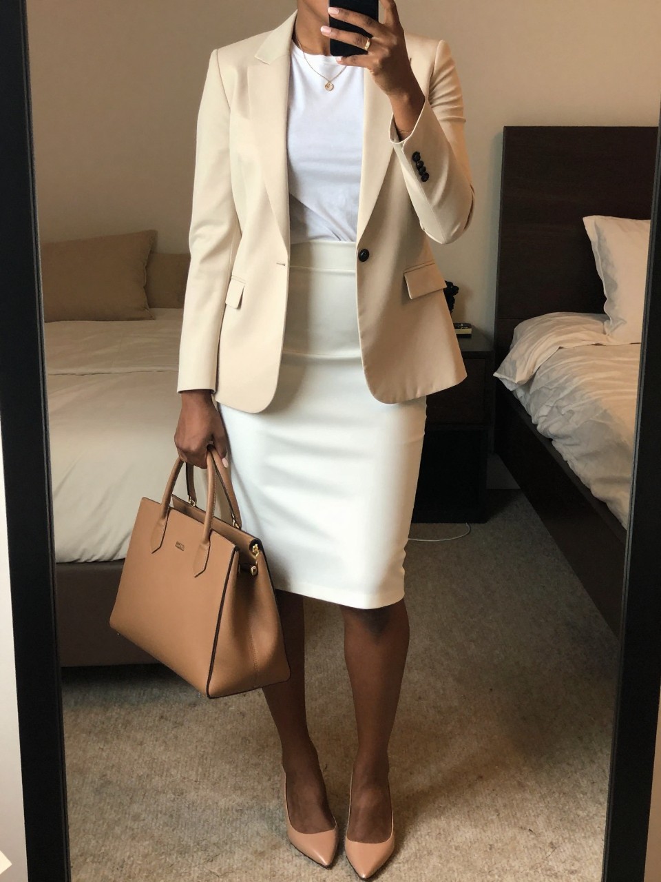 mirror selfie of a black woman wearing a cream blazer over a white tee tucked into a white pencil skirt with nude pointed flats, holding a structured tan bag, standing in a professional pose, cropped at NECK so face doesn't show, in a feminine-themed bedroom, warm moody lighting, iPhone photo quality.