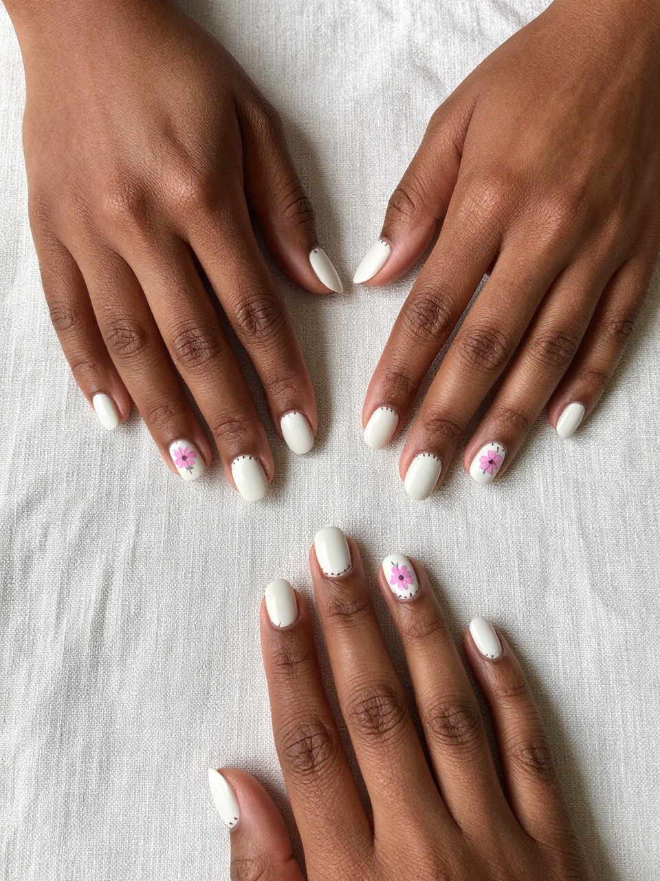 Top-down view of black feminine hands with short nails resting on a textured linen tablecloth. The nails are a matte off-white. Small dusty pink flowers are painted with tiny brown "stitch" marks around the edges of the nail design. The vibe is crafty, cozy, and handmade.