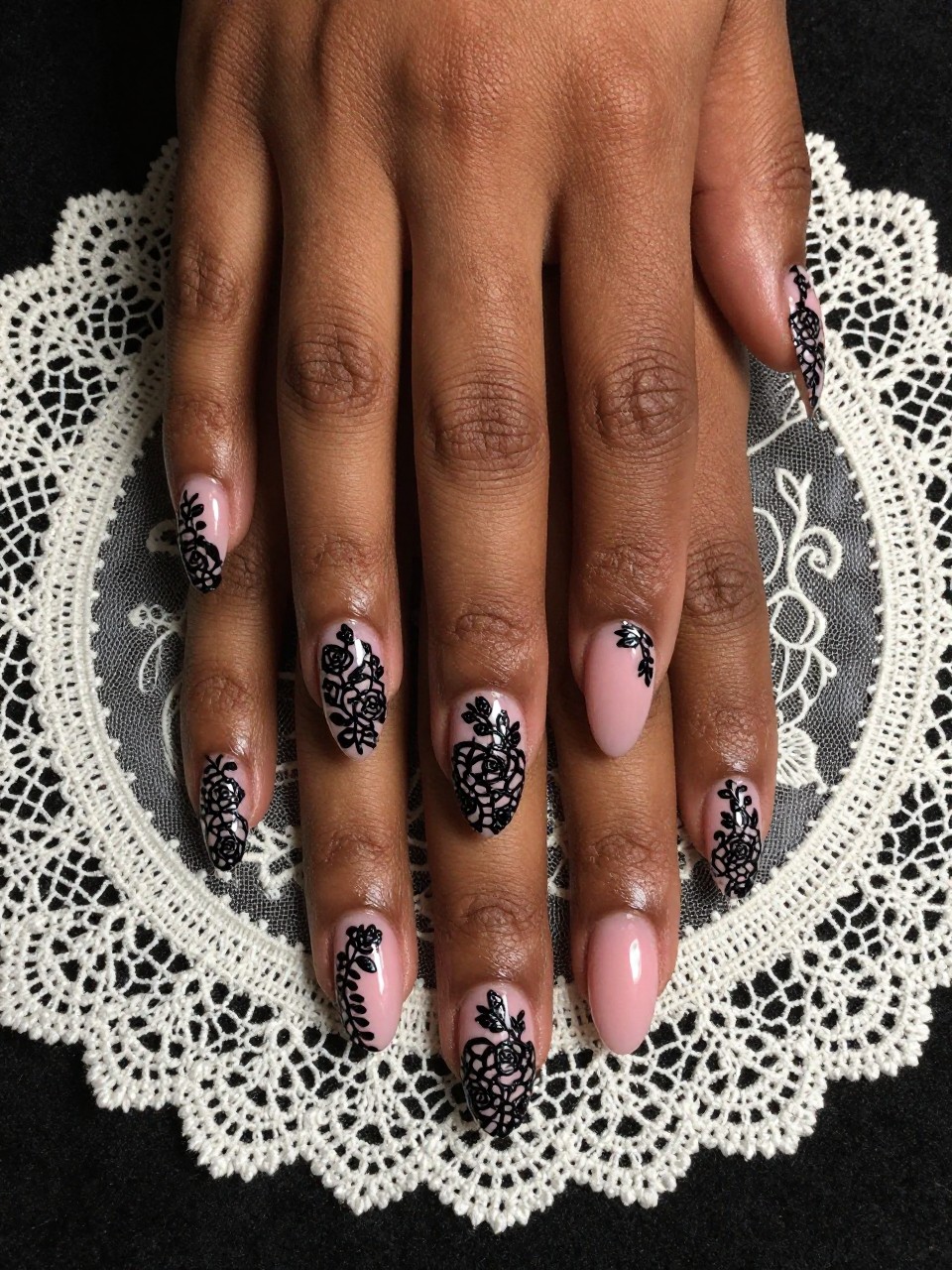 Bird's-eye view of black feminine hands with long, oval nails resting on an antique lace collar. The nails have a sheer pink base. An elaborate, hand-painted black lace pattern with small rose shapes covers each nail. The lighting is soft and slightly dramatic.