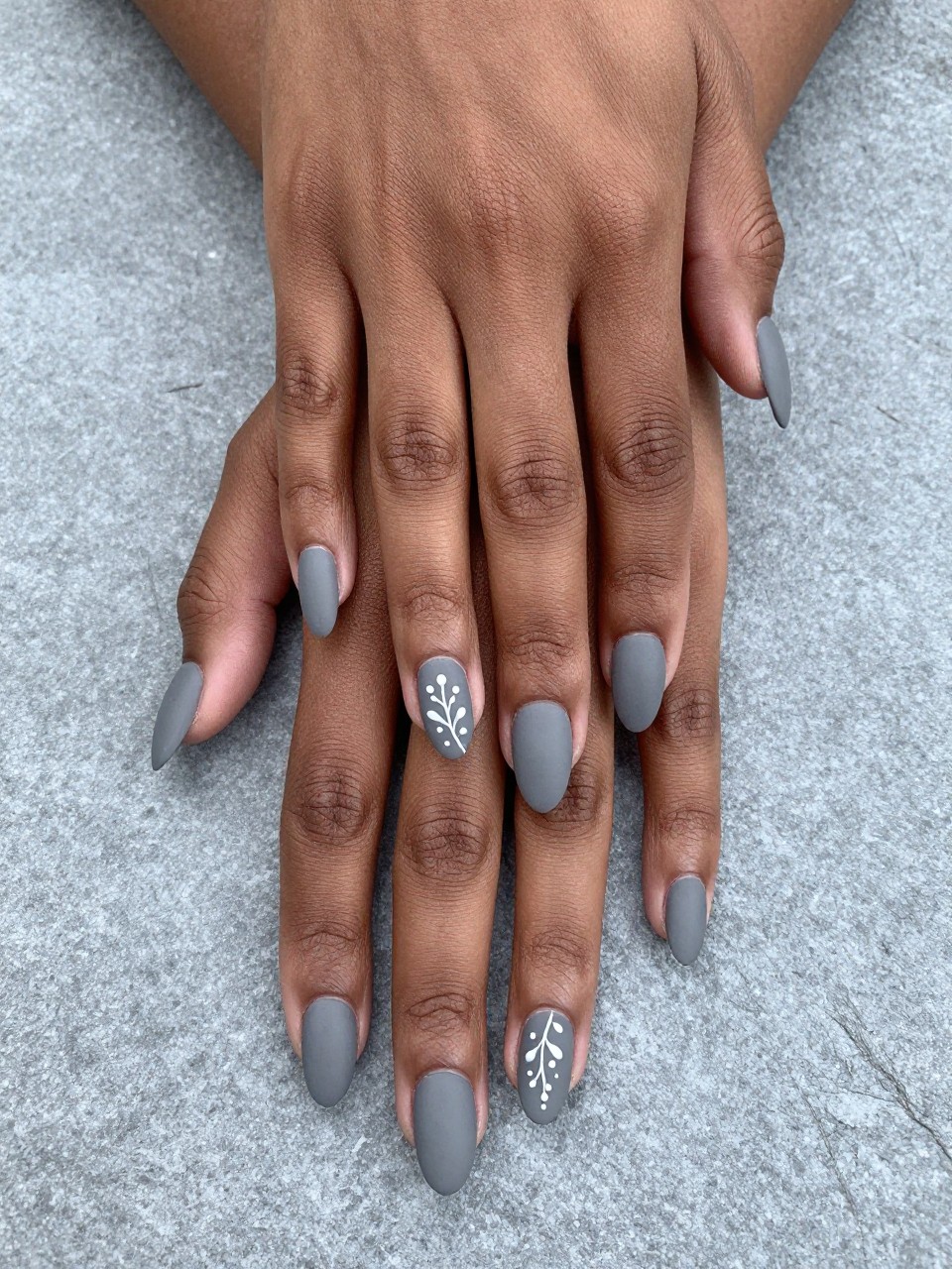 Top-down view of black feminine hands with almond nails on a cool grey stone surface. The nails are a matte heather grey. A single, fine white line forms a delicate vine with micro leaves and dots across each nail. The aesthetic is modern, minimalist, and artistic.