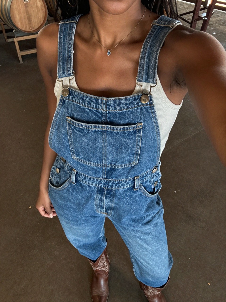 selfie of a black woman in denim overalls and a tank top with cowboy boots, standing at a farm or outdoor venue, cropped at NECK so face doesn't show, at a winery, warm moody lighting, iPhone photo quality.