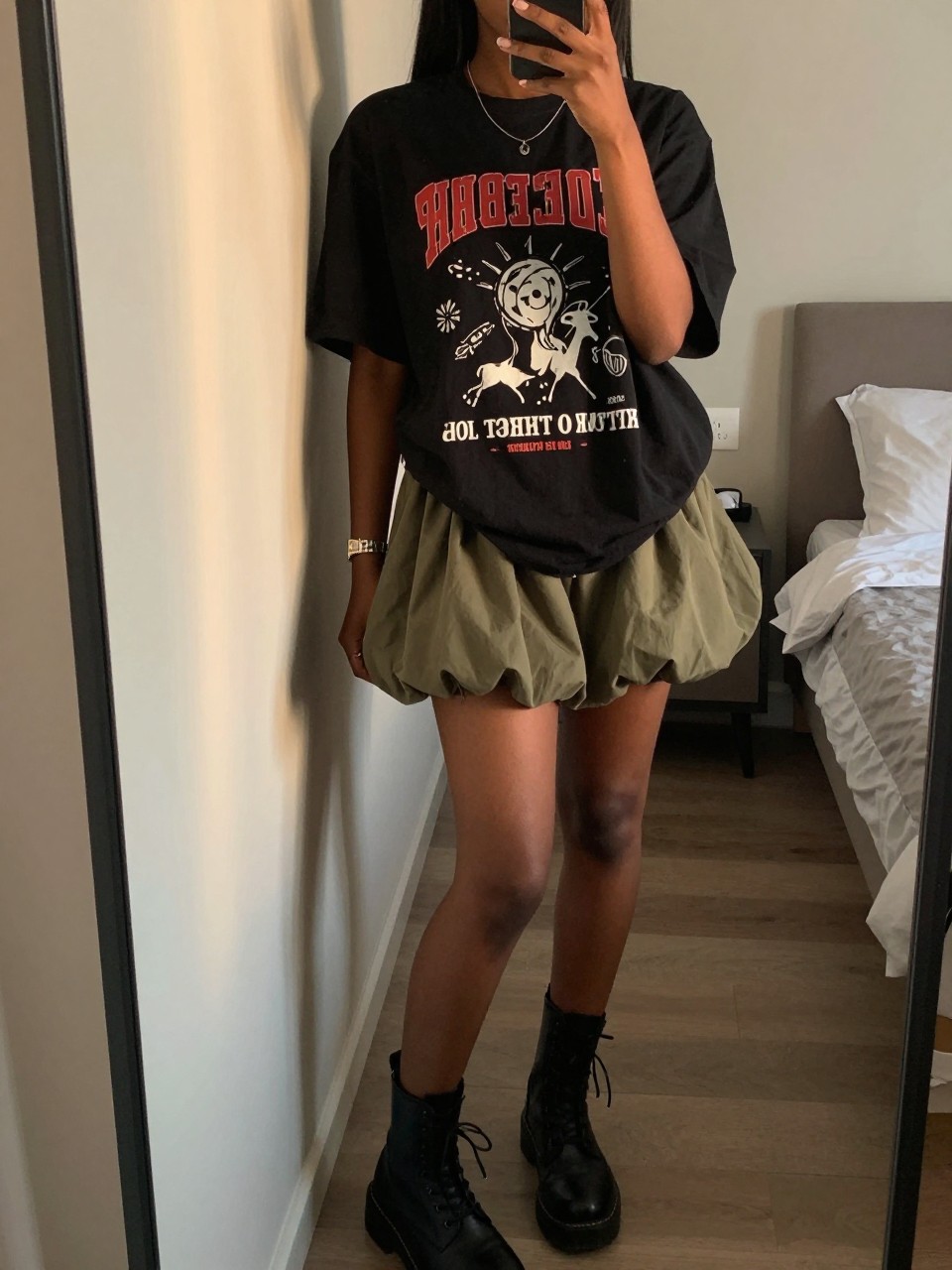 iPhone mirror selfie of a black woman in an oversized black graphic tee and an olive green bubble skirt with combat boots, casually leaning against a wall, cropped at NECK so face doesn't show, in a feminine-themed bedroom, evening golden hour, iPhone photo quality.