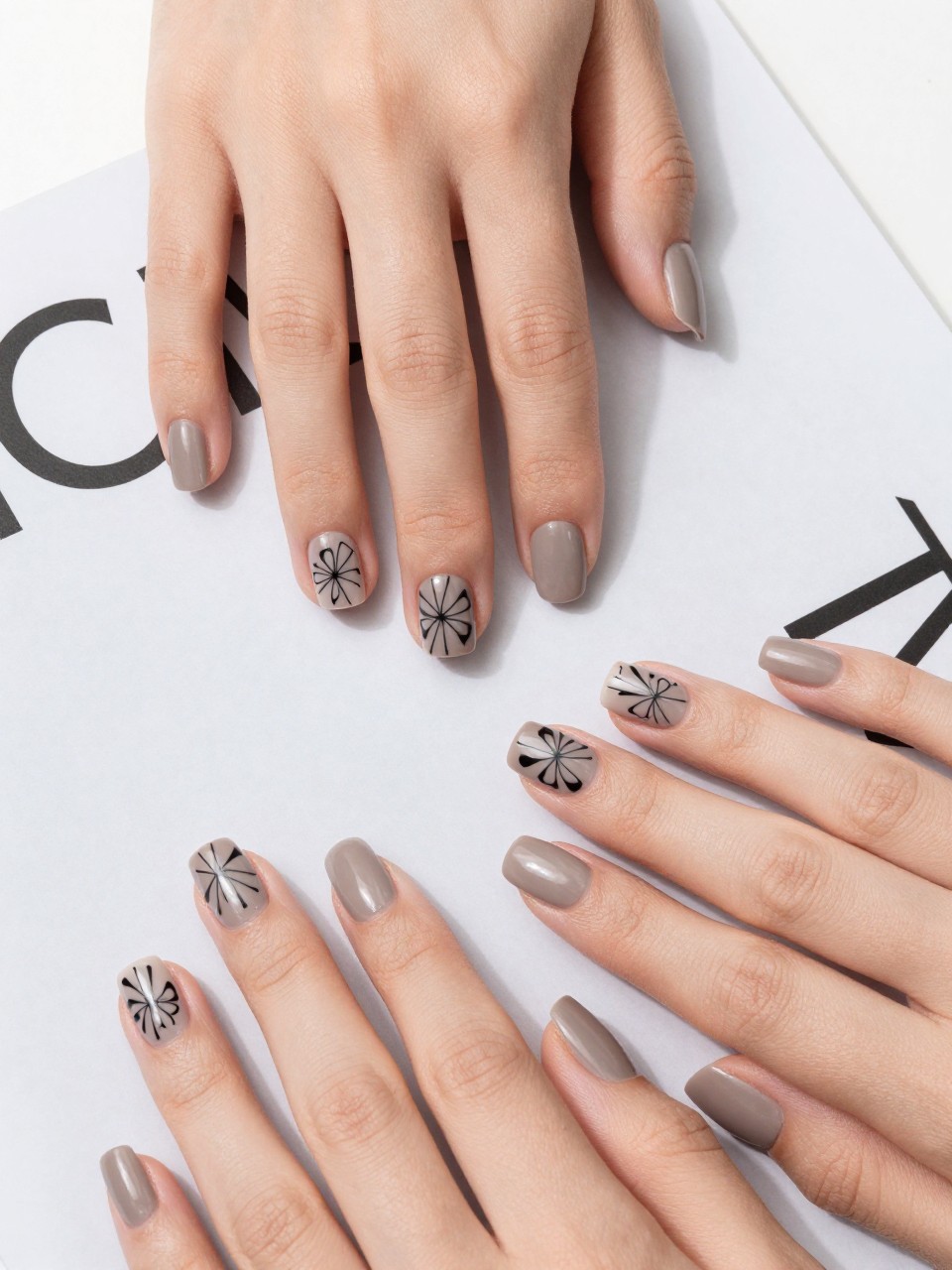 Overhead view of hands with short, square nails on a modern design magazine. The nails are a matte taupe and on each nail, there is a floral design that is composed of clean black geometric shapes forming abstract flowers. The setting is a bright, minimalist studio.