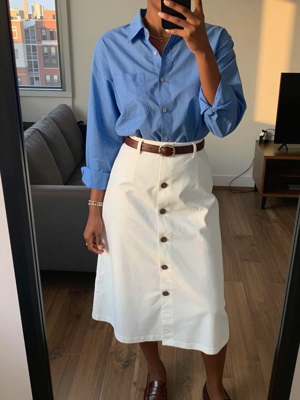 mirror selfie of a black woman wearing a blue oxford shirt tucked into a white button-front skirt with a brown leather belt and loafers, one hand adjusting her sunglasses on her head, cropped at NECK so face doesn't show, in a city apartment, evening golden hour, iPhone photo quality.