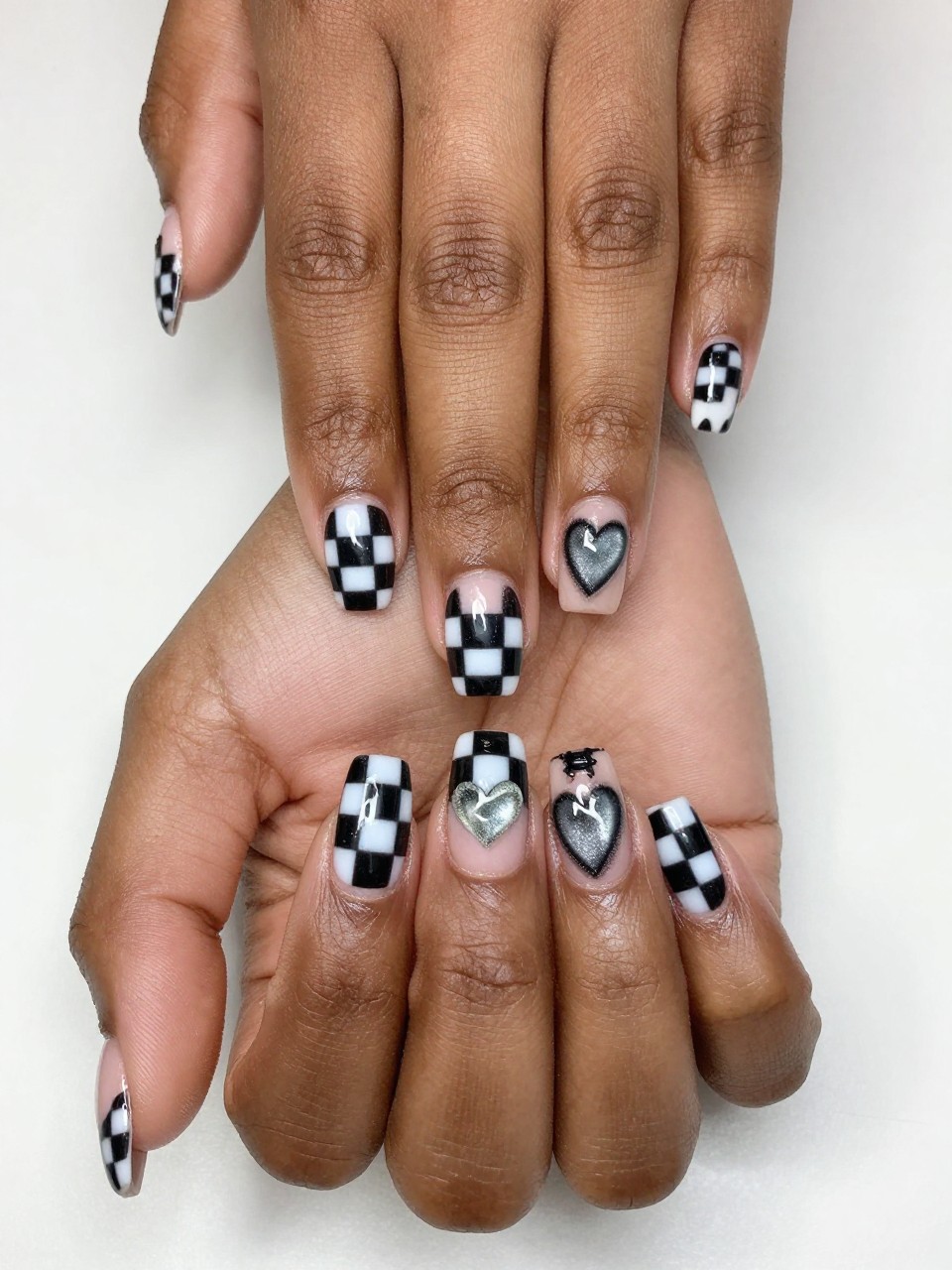 Top-down view of black feminine hands with a mix of checkered nails and one accent nail. The accent nail has a checkered pattern with one square replaced by a shimmering cat eye heart. The design is clever and fun.