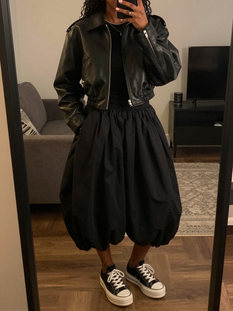iPhone mirror selfie of a black woman in a cropped black leather jacket and a black bubble skirt with platform sneakers, one hand in her jacket pocket, cropped at NECK so face doesn't show, in a city apartment, warm moody lighting, iPhone photo quality.