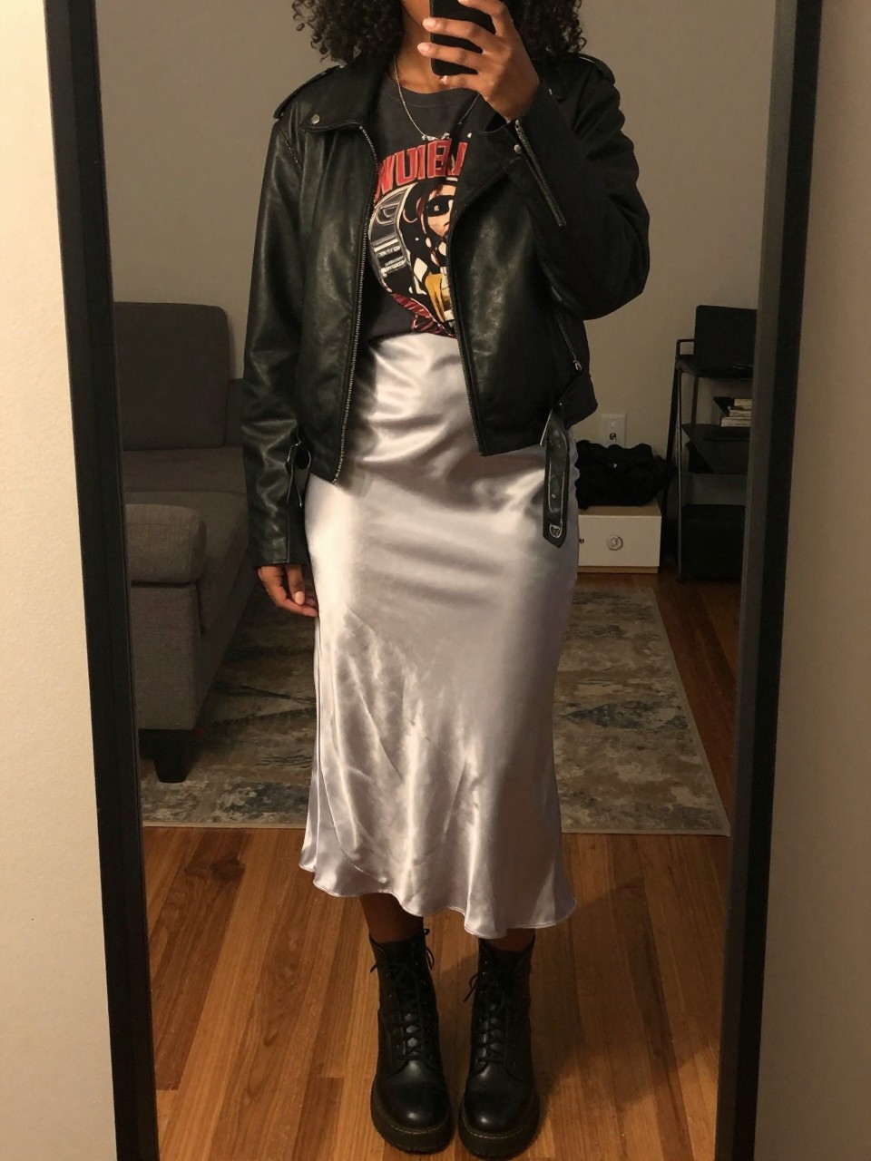 iPhone mirror selfie of a black woman wearing a black leather jacket over a vintage band tee with a silver satin midi skirt and black combat boots, one hand in her jacket pocket, cropped at NECK so face doesn't show, in a city apartment, warm moody lighting, iPhone photo quality.