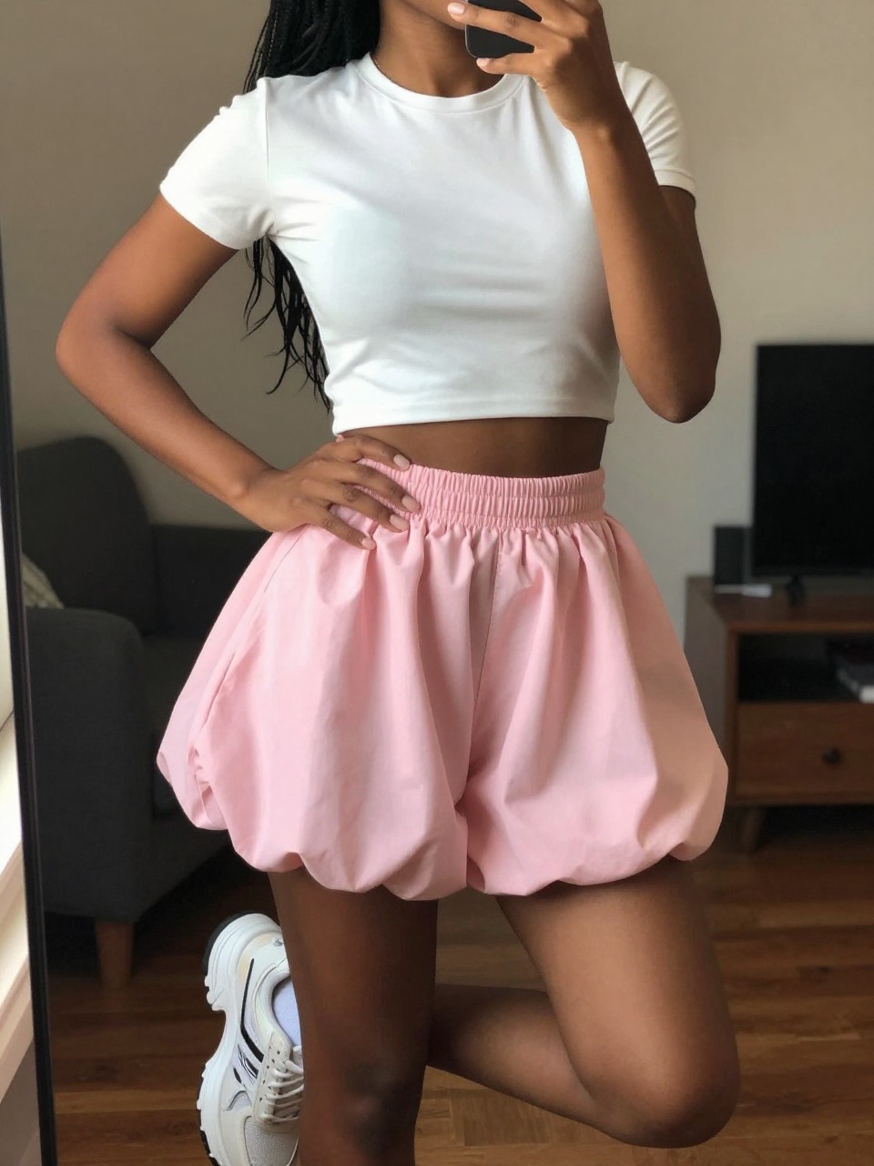 iPhone mirror selfie of a black woman in a white cropped baby tee and a pastel pink bubble skirt with chunky sneakers, one hand on her hip, cropped at NECK so face doesn't show, in a city apartment, warm moody lighting, iPhone photo quality.