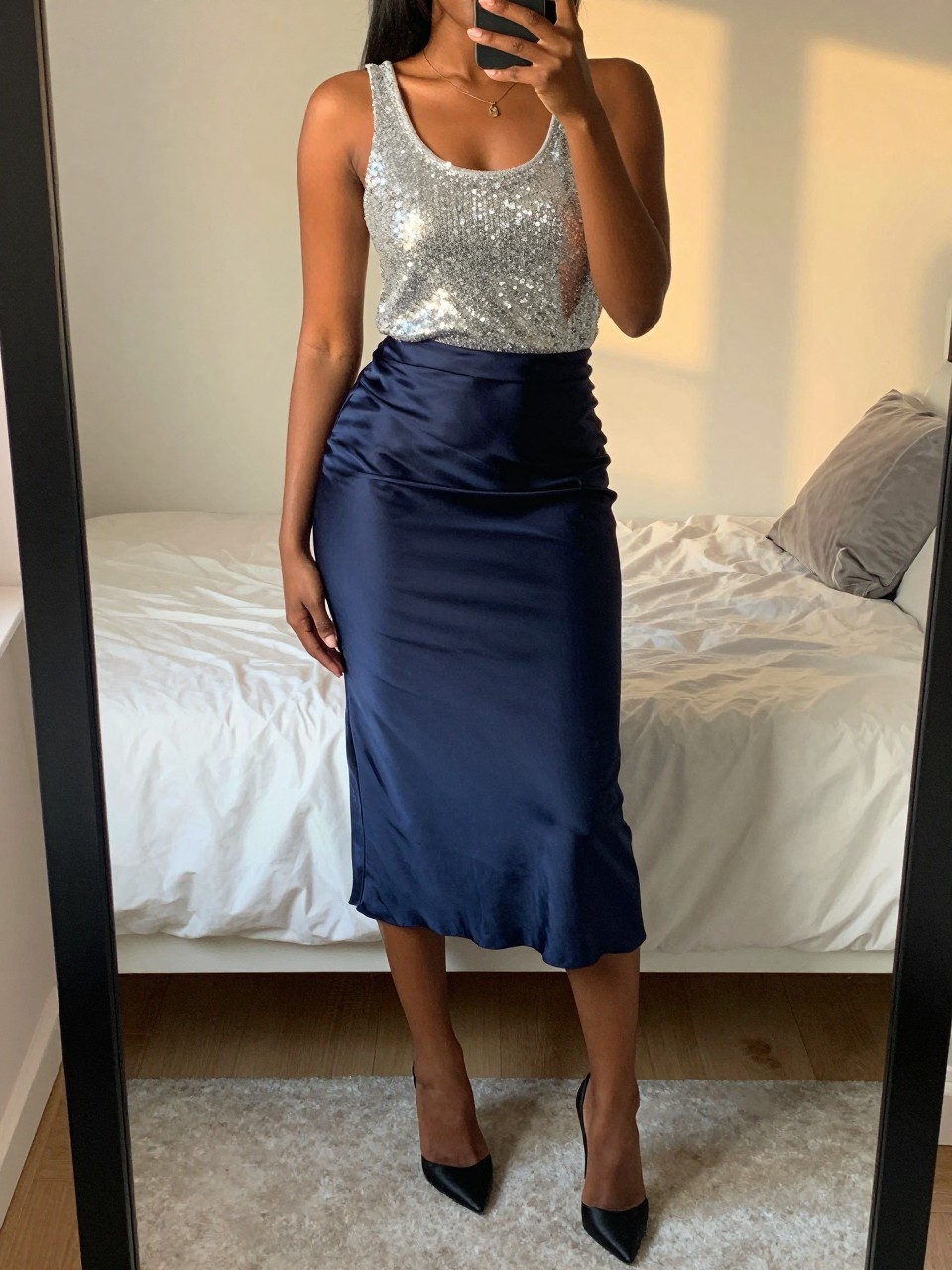 iPhone mirror selfie of a black woman wearing a silver sequin tank top tucked into a navy satin midi skirt with black satin stiletto heels, one hand holding her phone at her side, cropped at NECK so face doesn't show, in a feminine-themed bedroom, evening golden hour, iPhone photo quality.